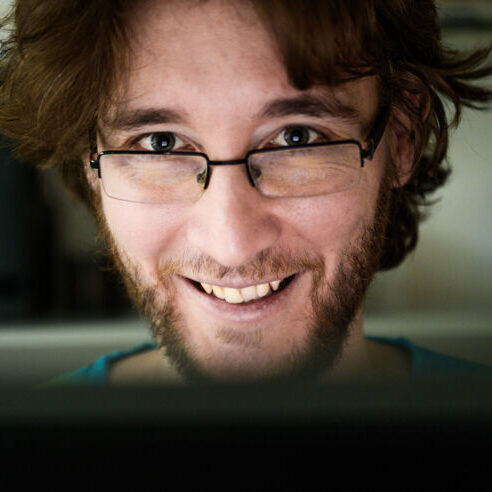 A young man with medium-length brown hair, glasses, and a short beard smiles warmly while looking at a computer screen, with a blurred background behind him.