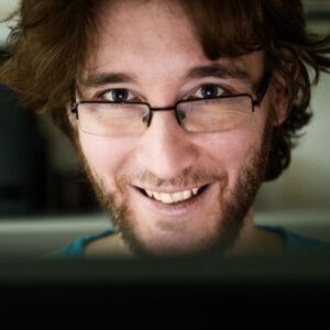 A person with glasses and a beard smiles warmly at the camera, sitting indoors with a blurred background.