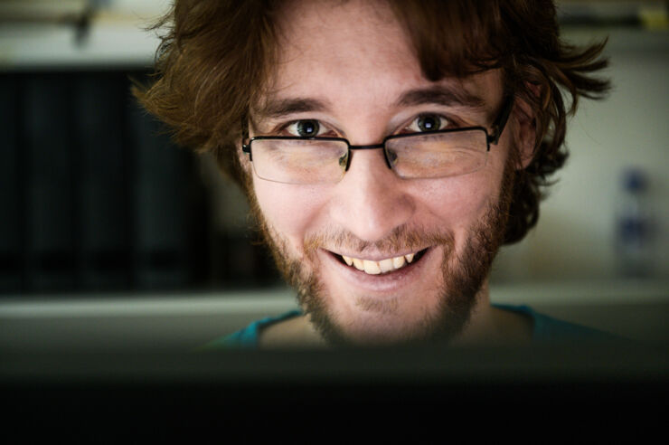A man with glasses and a beard smiles whilst looking at a computer screen, with soft lighting and shelves blurred in the background.