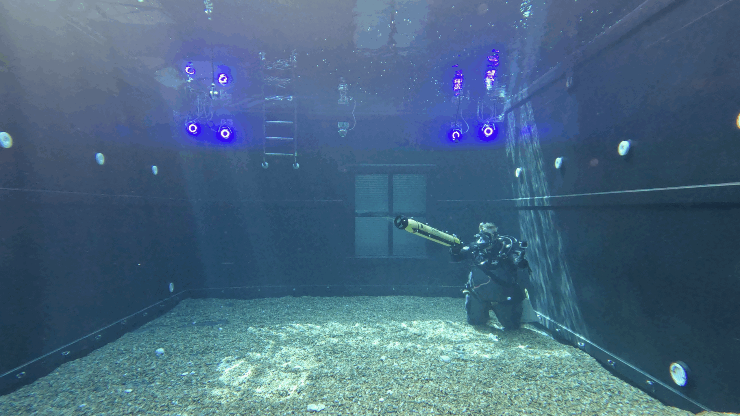 A scuba diver kneels on the gravel floor of a large, dark tank, holding a yellow underwater device. Blue lights and cameras are mounted on the tank’s walls and ceiling, and a ladder is visible at the back.