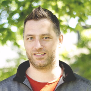 A man with short dark hair and light facial hair is standing outdoors in front of leafy green trees, wearing a grey zip-up jacket over a red shirt, and smiling slightly at the camera.