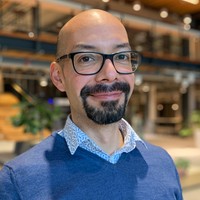 A person with a shaven head, glasses, and a short beard smiles whilst standing indoors in a modern, open space with wood and metal accents. They are wearing a blue jumper over a patterned collared shirt.