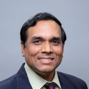 A man with short dark hair wearing a dark suit, light green striped shirt, and maroon tie, smiling in front of a plain grey background.