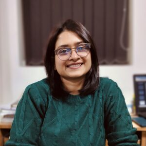 A woman with shoulder-length dark hair and glasses, wearing a green jumper, smiles whilst sitting indoors at a desk with books and a laptop in the background.