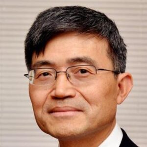 A man with short, dark hair and glasses, wearing a suit and white shirt, smiles slightly in front of a neutral background with horizontal lines.