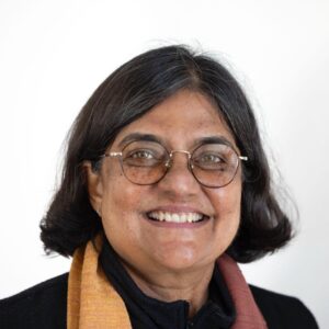 A woman with short dark hair and glasses smiles at the camera. She is wearing a black top and an orange scarf, with a light-coloured background.