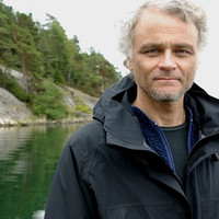 A man with curly grey hair and a short beard stands by a calm lake, wearing a black jacket. Trees and rocky cliffs are visible in the background under an overcast sky.