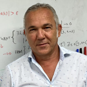 A man with short grey hair, wearing a light-coloured, patterned dress shirt, stands in front of a whiteboard covered with mathematical equations and notes written in various colours.