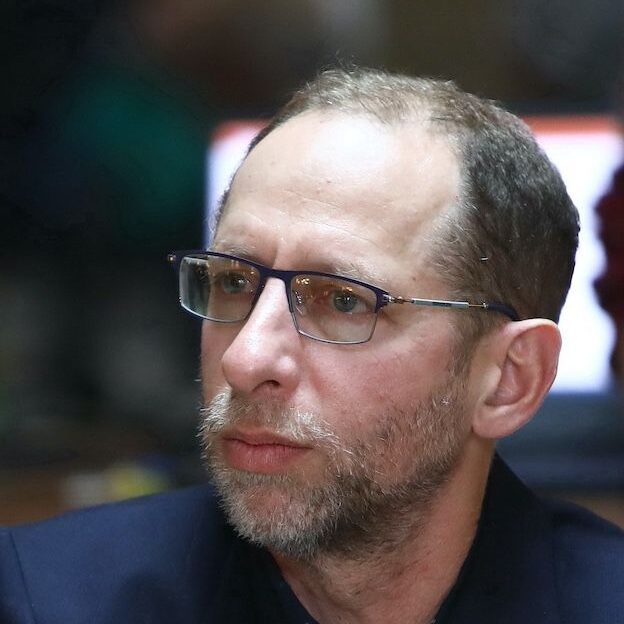 A middle-aged man with short brown hair, glasses, and a trimmed beard looks focused. He is wearing a dark jacket and is sitting indoors with a blurred background.