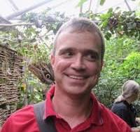 A man wearing a red shirt and carrying a shoulder bag smiles whilst standing in a lush, green indoor garden with sunlight filtering through a glass ceiling. Another person is visible in the background.