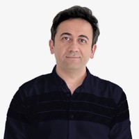A man with short dark hair wearing a dark button-up shirt stands facing the camera against a plain white background.