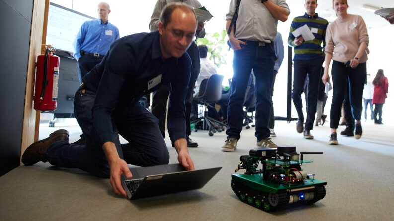A man kneels on the floor using a laptop to control a small, green robotic vehicle whilst several people stand and watch in a brightly lit indoor space.