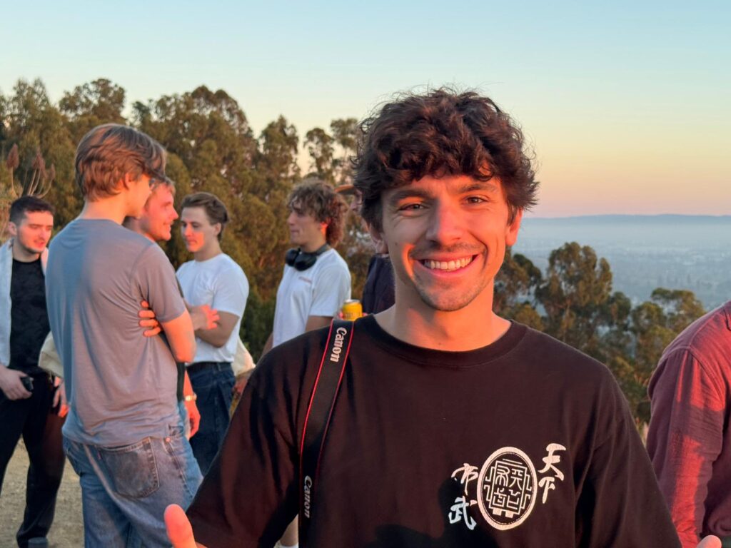 A smiling young man gives a thumbs up at sunset, standing outdoors with a camera strap over his shoulder. Several people are chatting in the background, with trees and a hazy sky visible behind them.