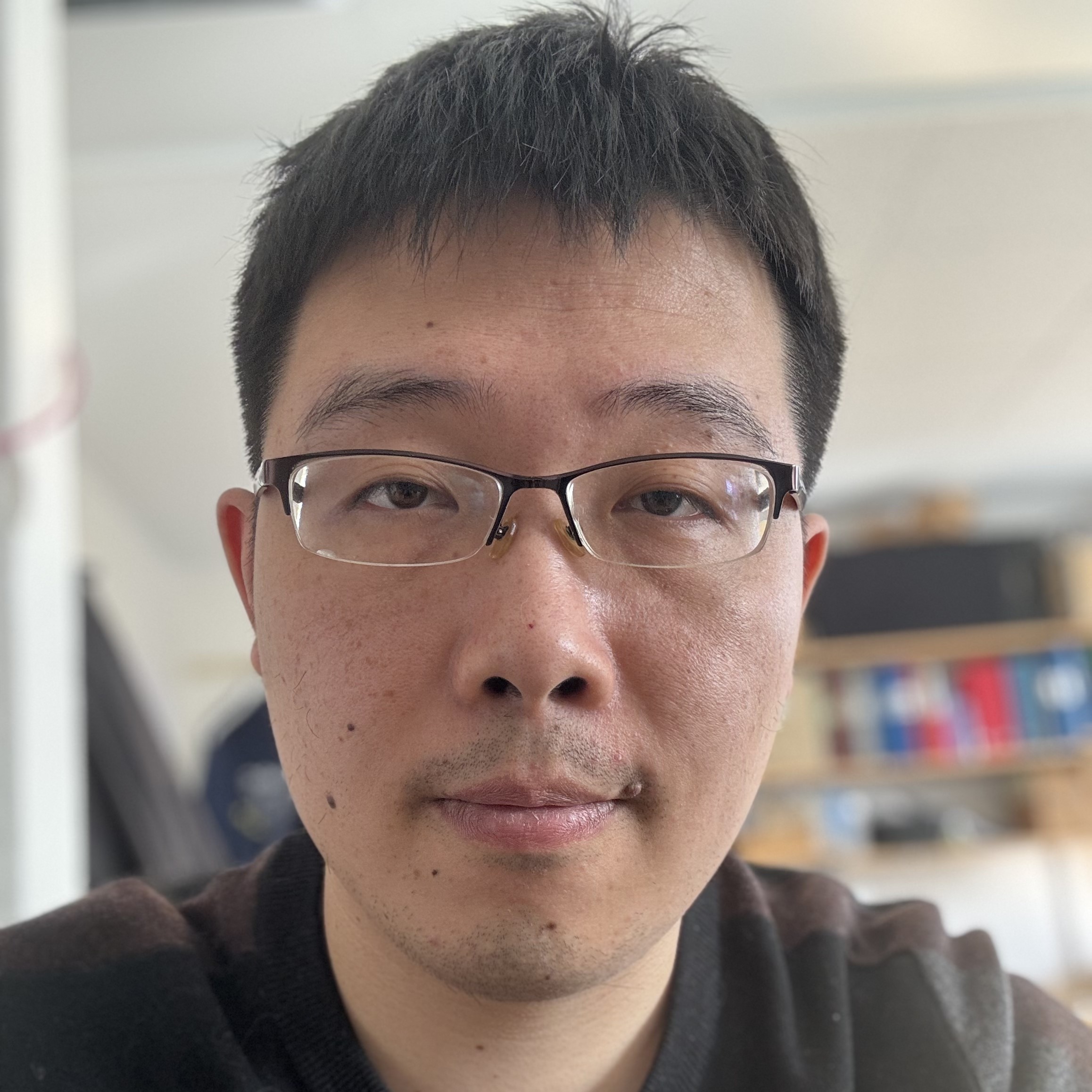 A man with short black hair and glasses looks directly at the camera, indoors with shelves and various items blurred in the background. He is wearing a brown and black jumper.