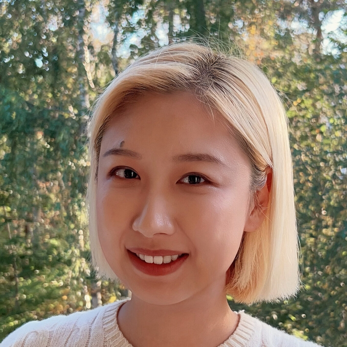 A person with short blonde hair smiles at the camera, wearing a cream-coloured cable-knit jumper. Green trees and sunlight are visible in the blurred background.