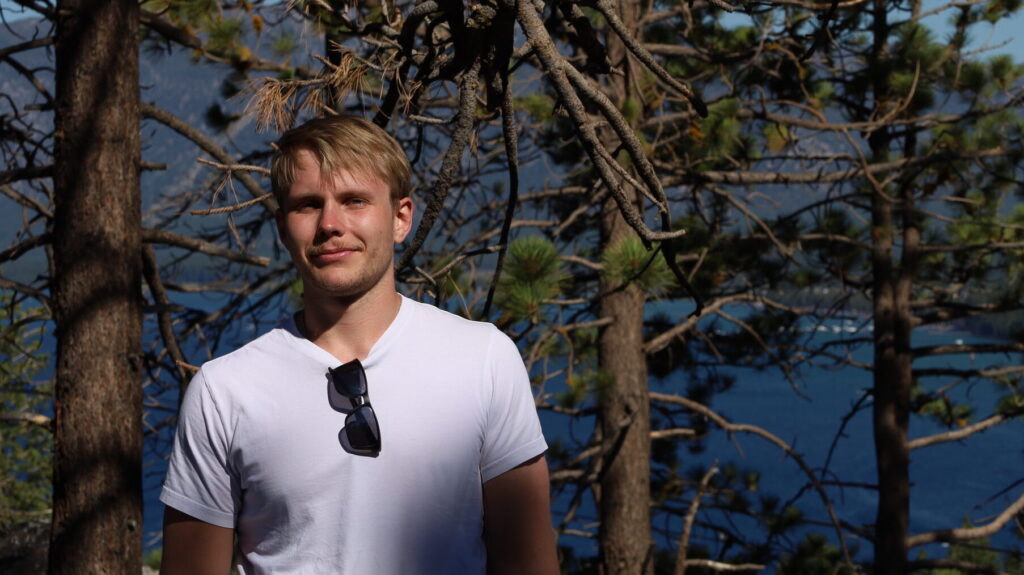 A man in a white T-shirt stands in front of pine trees with sunlight filtering through. Blue water and distant mountains are visible in the background, suggesting a scenic outdoor location. Sunglasses hang from his shirt.