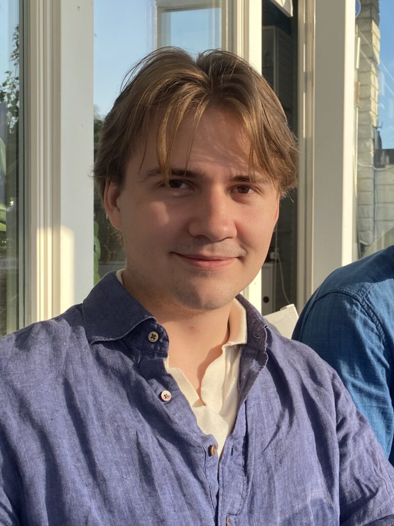 A young person with light brown hair and a slight smile sits indoors, wearing a blue button-up shirt over a white top. Sunlight is shining on their face and shoulder.