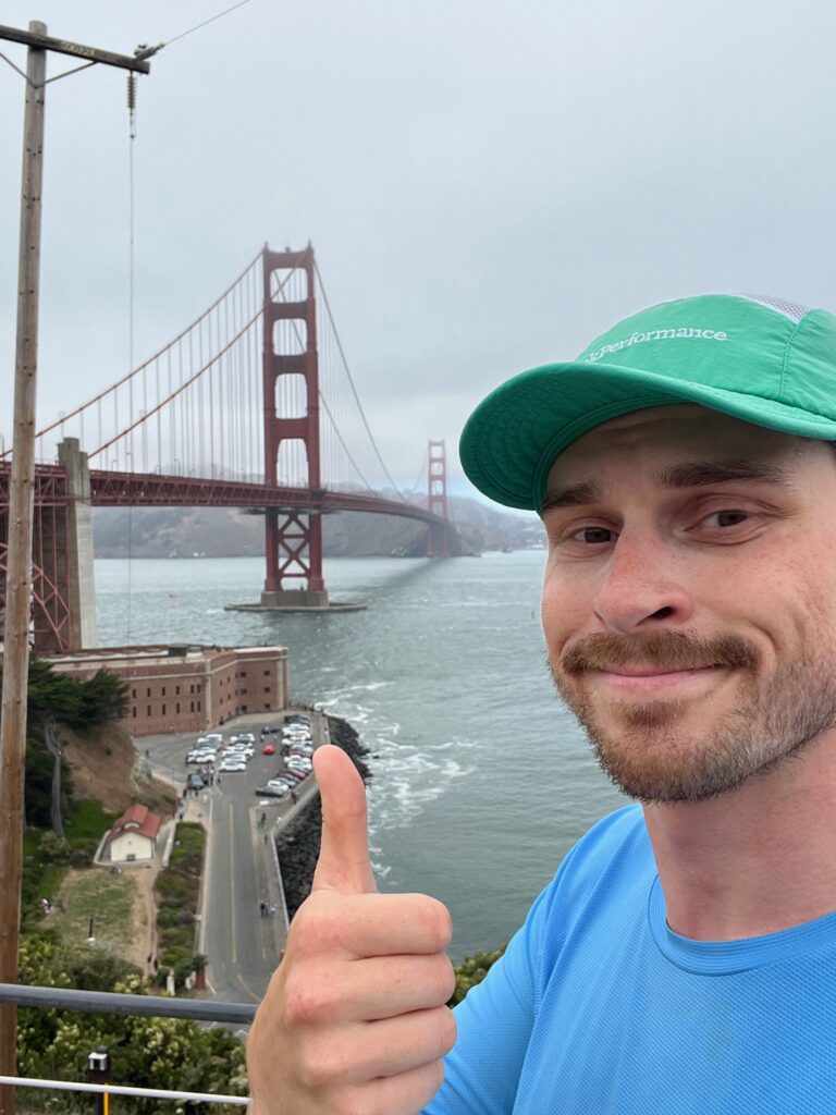 A man in a green cap and blue shirt gives a thumbs up and smiles at the camera with the Golden Gate Bridge in the background on a cloudy day.