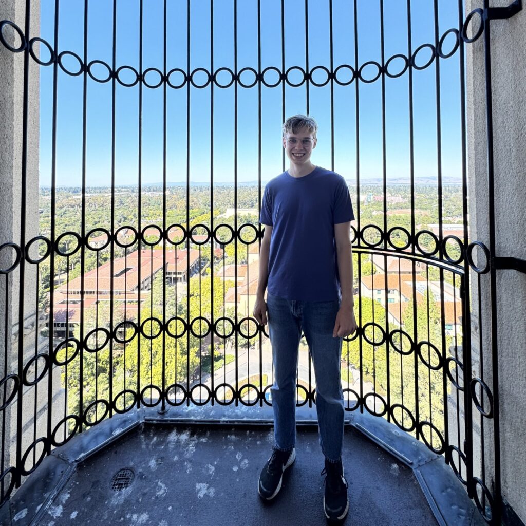 A young person in a blue shirt and jeans stands in front of a tall, decorative metal railing with a cityscape and clear blue sky visible in the background.