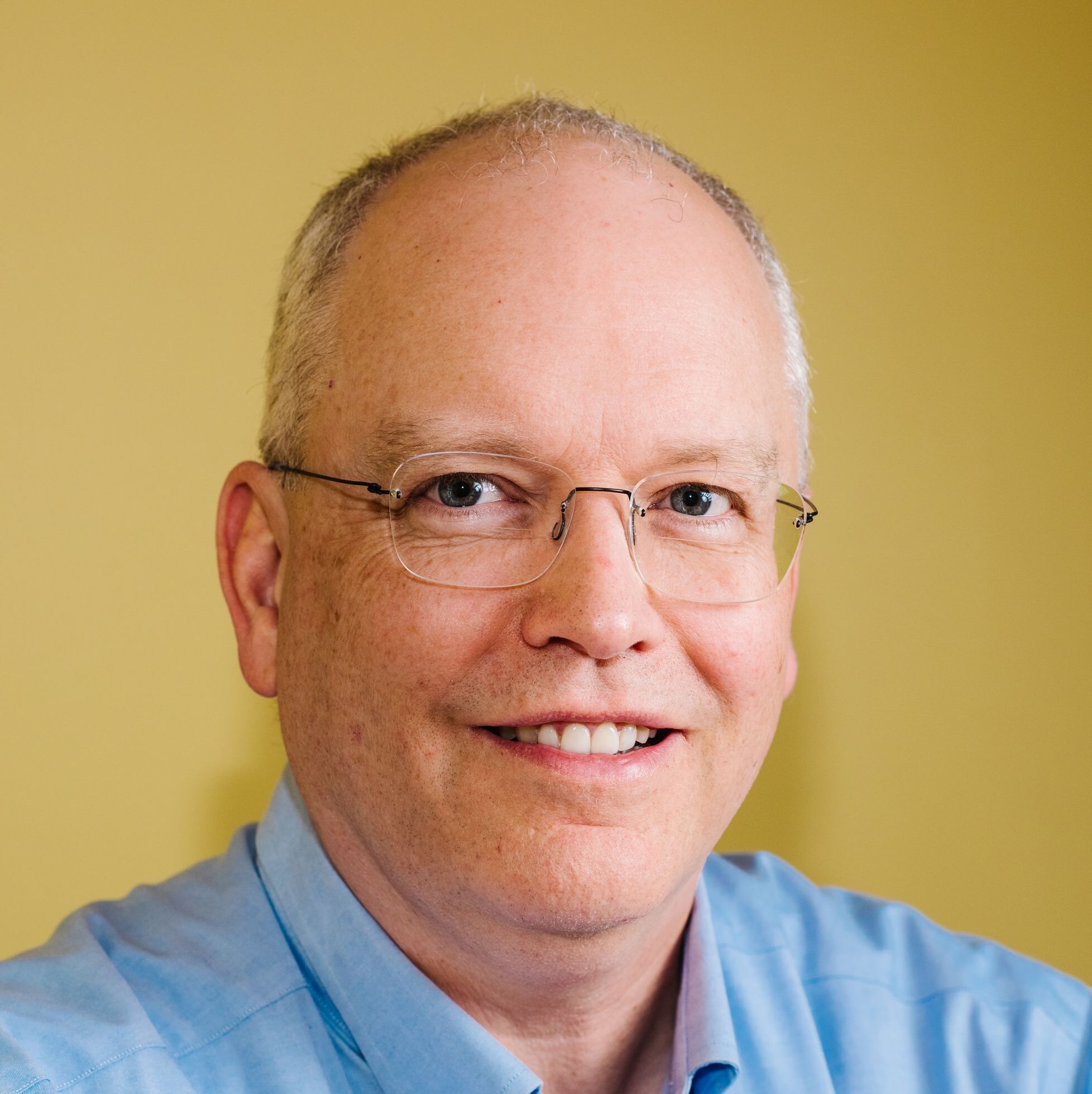 A middle-aged man with short grey hair and glasses, wearing a light blue collared shirt, smiling in front of a plain yellow background.