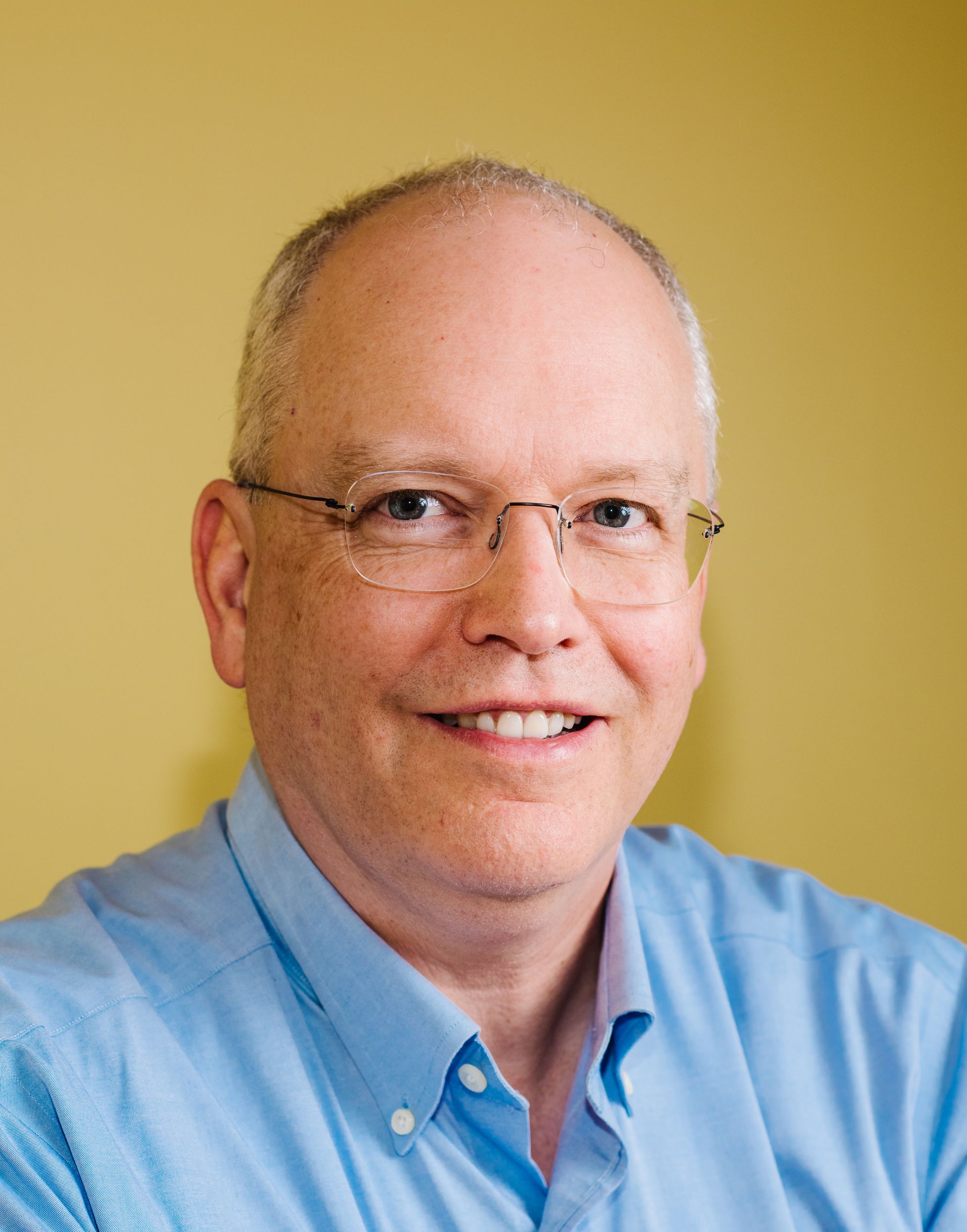 A middle-aged man with short grey hair and glasses, wearing a light blue collared shirt, smiling in front of a plain yellow background.