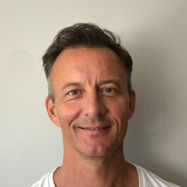 A middle-aged man with short brown hair and light stubble is smiling at the camera. He is wearing a white V-neck T-shirt and standing in front of a plain light-coloured wall.