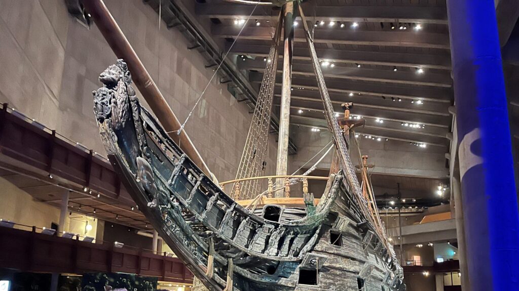 A large, historic wooden ship is displayed indoors at a museum-like venue, surrounded by people seated at dinner tables under dim lighting.