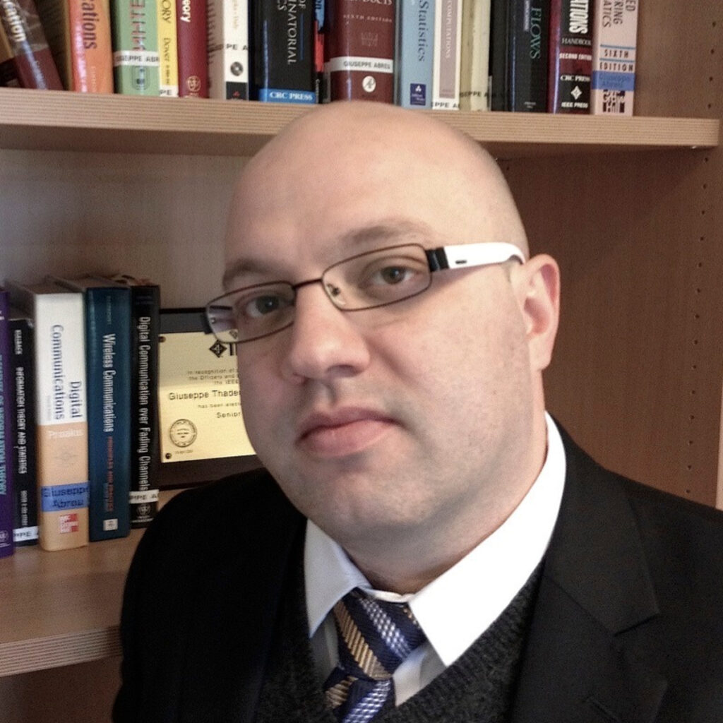 A bald man wearing glasses, a suit, and a tie is standing in front of a bookcase filled with various books. He is looking directly at the camera with a neutral expression.
