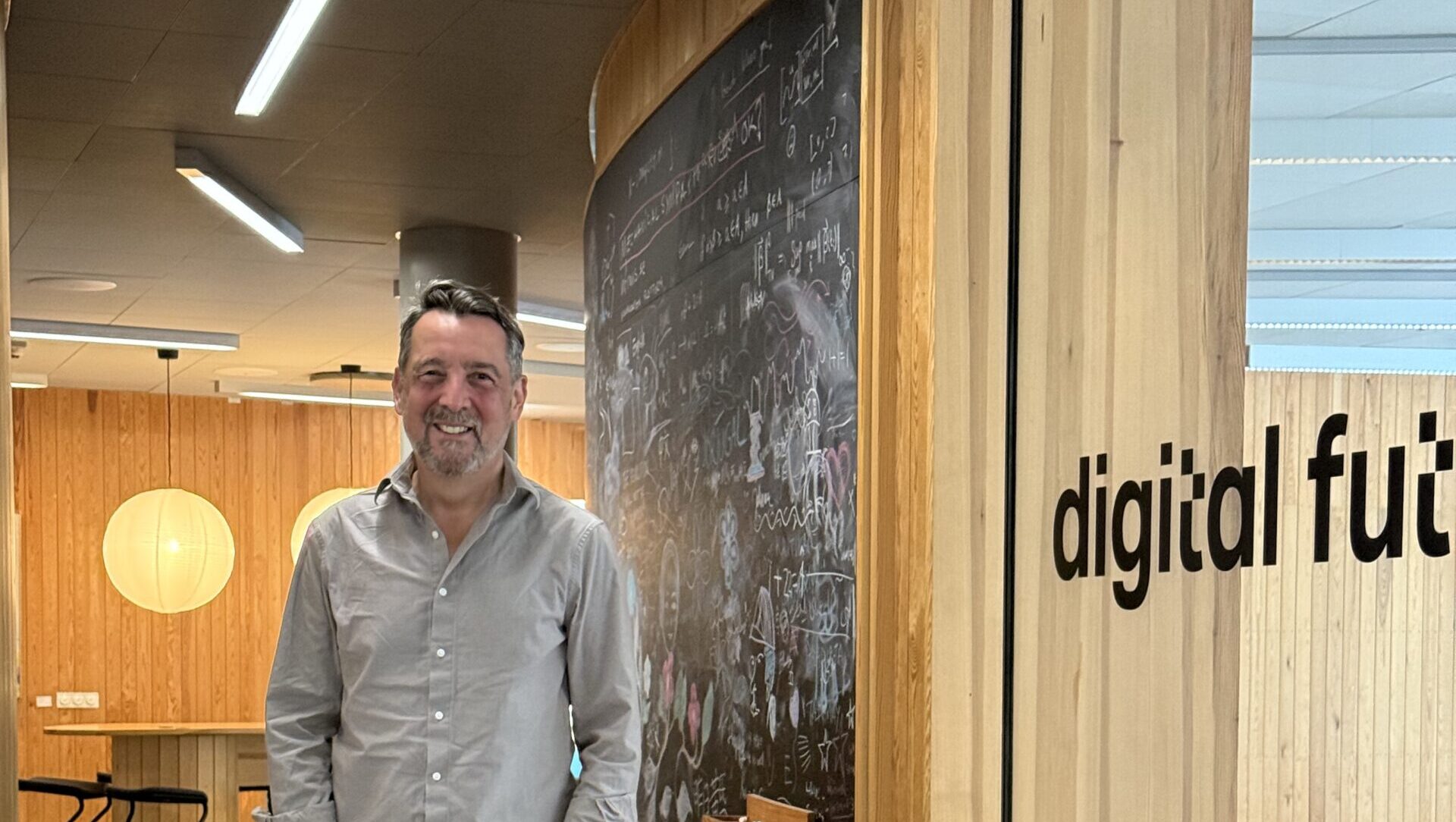 A man in a grey shirt and jeans stands smiling in a modern office corridor with wooden walls, a blackboard covered in writing, and a window labelled digital fut.