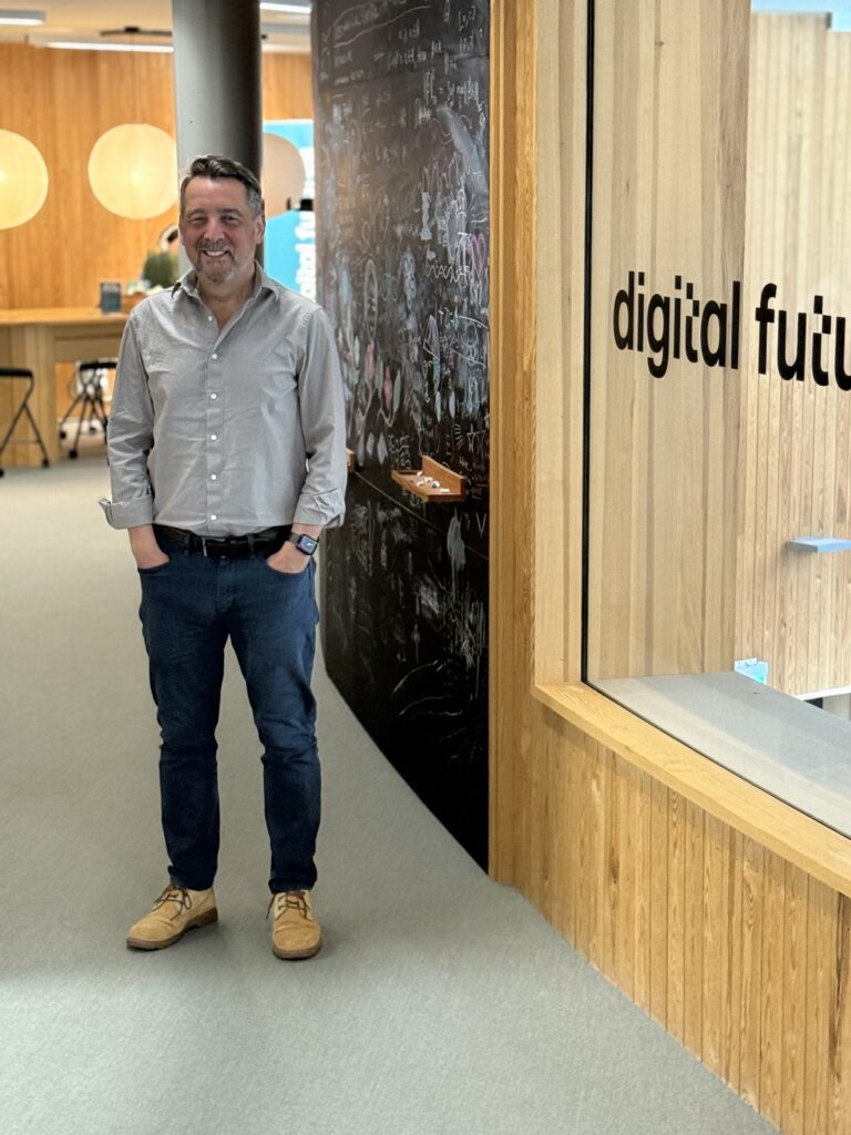 A man in a grey shirt and blue trousers stands smiling in a modern office space with wooden walls. Behind him is a wall with chalk writing and a glass window with digital futu visible.