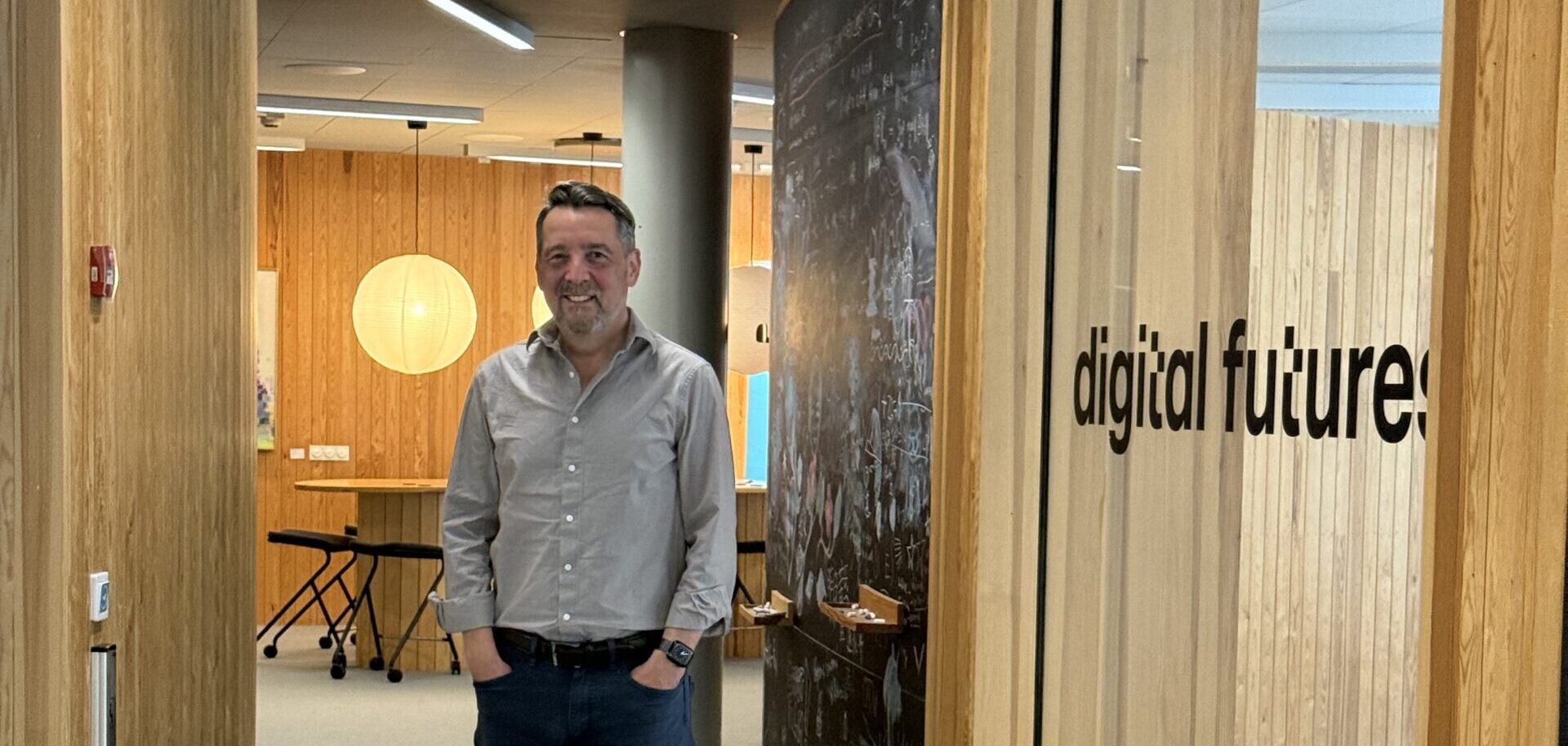 A man in a grey shirt and jeans stands in a modern office corridor with wooden walls. A window labelled “digital future.” is visible beside him. Overhead lights and an emergency exit sign illuminate the space.