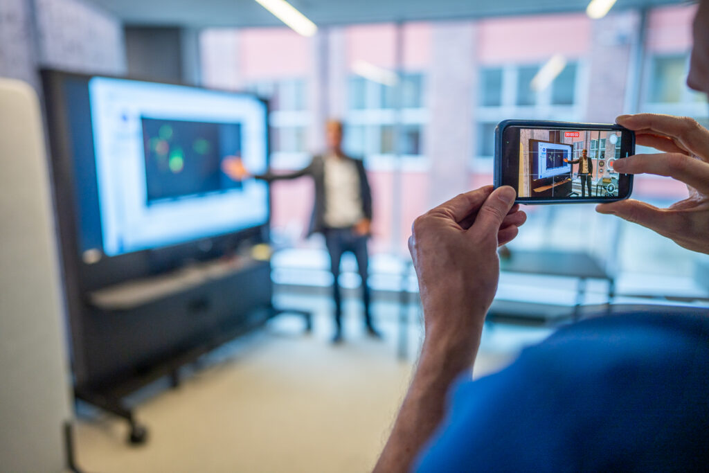 A person takes a photo on a mobile phone of a presenter standing by a large screen with a blurred digital display in a modern, bright office setting.