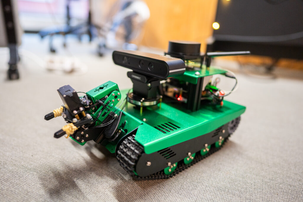A small green robot with tank tracks, a robotic arm, and sensors is positioned on a carpeted floor in an indoor setting.