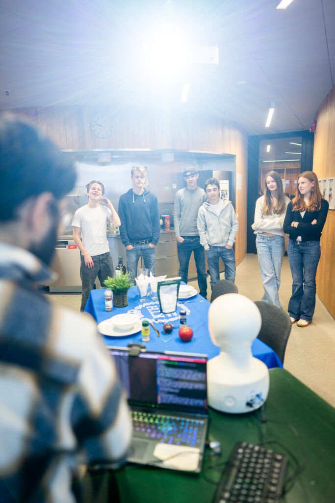 A group of six young people stand talking near a table set with crockery and robotics equipment, while a person in the foreground, partially out of focus, operates a laptop. Bright indoor lighting.