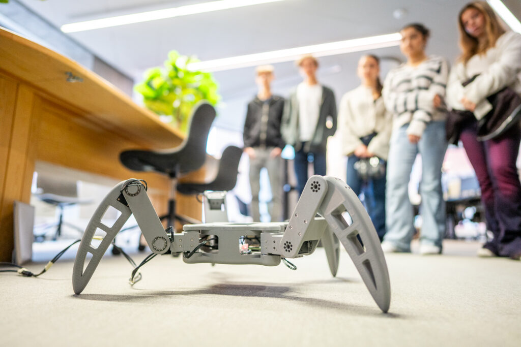 A group of people watches a grey, four-legged robotic device on the floor in an office or classroom setting with desks and bright lighting.