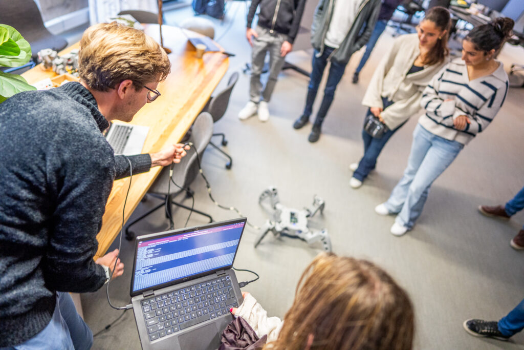 A group of people stands in a room watching a man operate a small quadruped robot using a laptop. The scene appears to be a workshop or demonstration, with desks and chairs in the background.