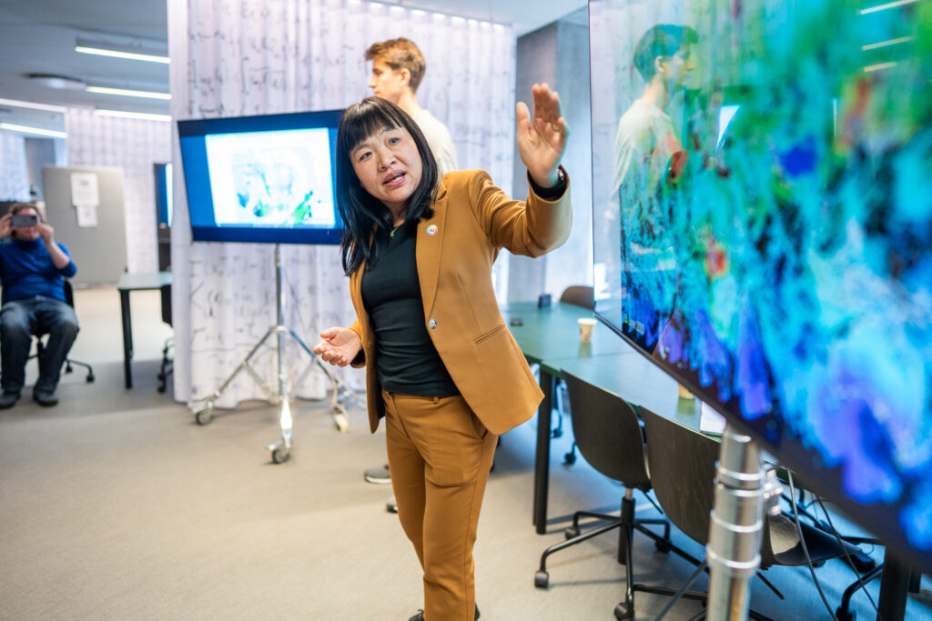 A woman in a brown suit gestures towards a colourful digital display in a modern office, whilst others watch and interact in the background.