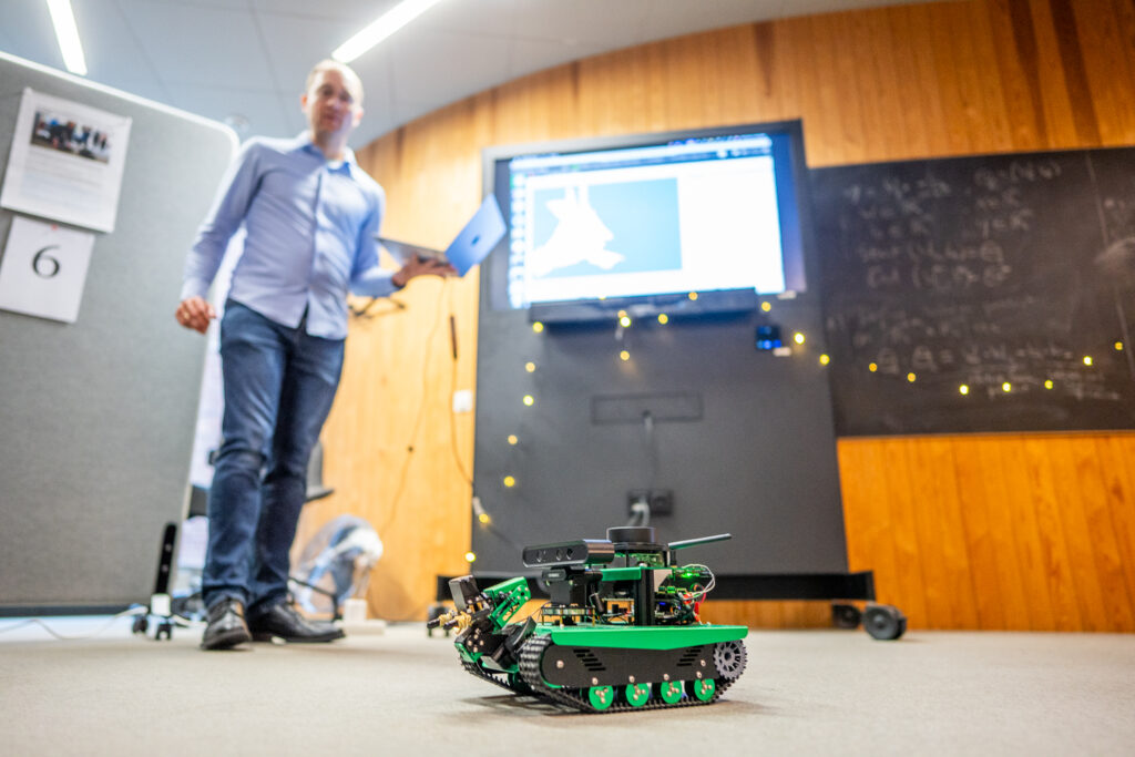 A small green robot tank moves on the floor in a classroom. In the background, a person stands holding a laptop near a screen displaying technical diagrams, with a blackboard and fairy lights on the wall.