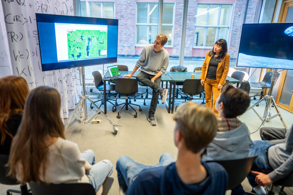 A man and a woman present data on a large screen to a seated group in a modern, bright classroom with large windows and mathematical equations on the walls.