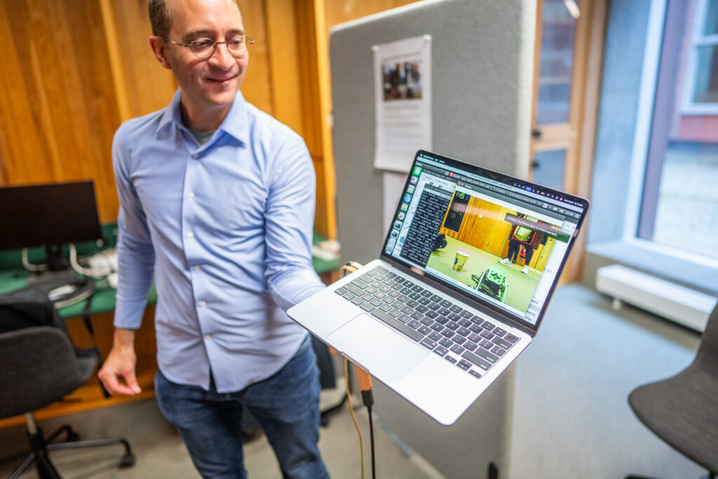 A man in glasses and a blue shirt holds a laptop displaying an image editing or video software interface in an office with wooden walls and computer desks.