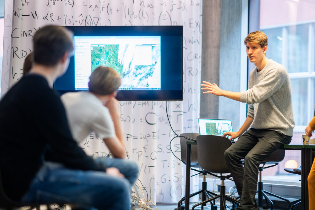 A man gestures while presenting satellite imagery on a screen to three seated people in a modern classroom with scientific equations written on a whiteboard behind them.