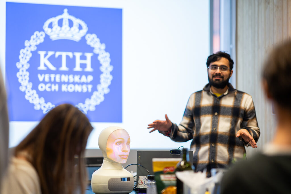 A man gives a presentation beside a mannequin head with a projected face, in front of a screen displaying the KTH logo and Swedish text “Vetenskap och Konst.” Several people are watching.