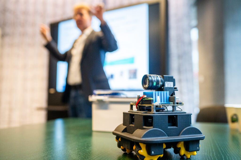 A small robotic vehicle with a camera and sensors sits on a table in focus, while a blurred person in business attire presents in the background near a large display screen.