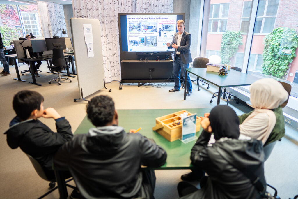 A person stands next to a large monitor giving a presentation to four seated people in a modern classroom. Mathematical equations are written on a glass wall in the background.
