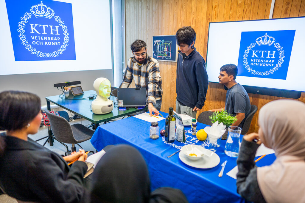 A group of students presents a project with a mannequin head and electronics at a table set with food items, in a classroom with KTH Royal Institute of Technology logos on screens and wood-panelled walls.