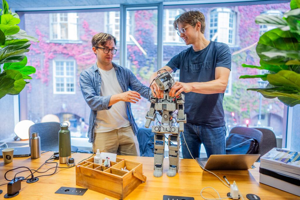 Two people stand at a table in a bright room, working together to adjust a humanoid robot. The table is covered with electronics, tools, coffee cups, and a laptop. Large windows and leafy plants surround the scene.
