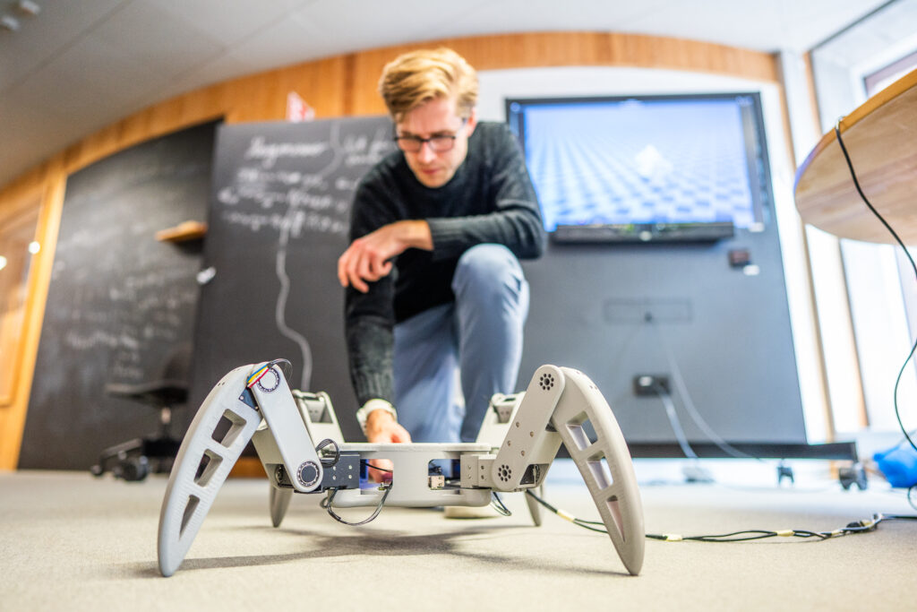 A person kneels on the floor, working on a white, four-legged robot in a modern room with a blackboard, TV screen, and wooden walls in the background.