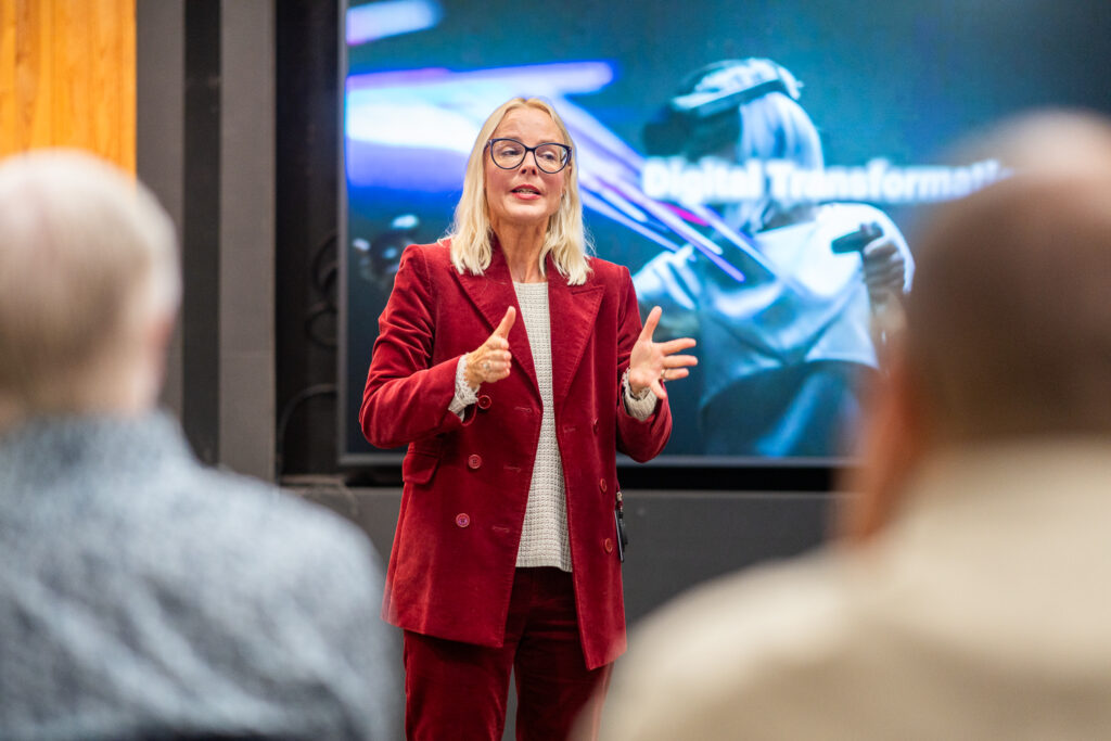 A woman in a red suit is speaking to an audience in front of a screen displaying the words Digital Transformation and an image of a person in a VR headset.