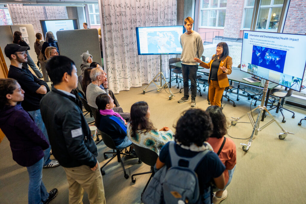 A woman presents to a group in a classroom with large monitors displaying maps and images. People are seated and standing, listening attentively. Scientific equations are visible on whiteboards in the background.