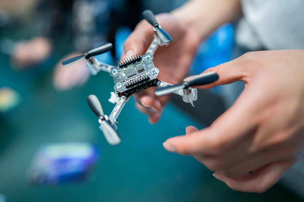A person holds a small quadcopter drone with exposed circuitry and four black propellers, focussing on the drone whilst the background is blurred.